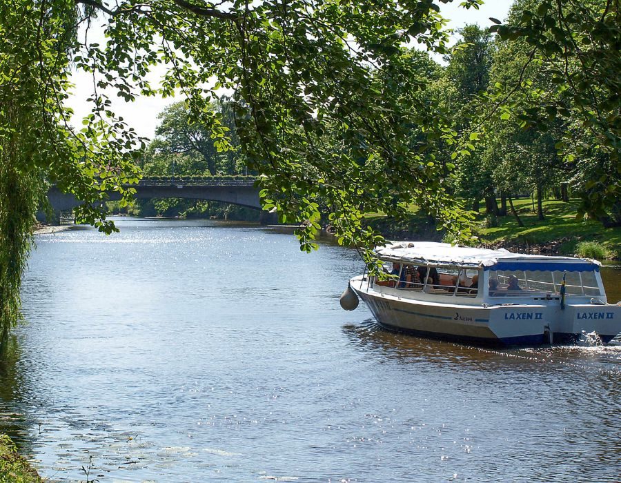 Turbåten Laxen på Rönne å i Ängelholm med grönskande natur och bro i bakgrunden nära Destination Råbocka